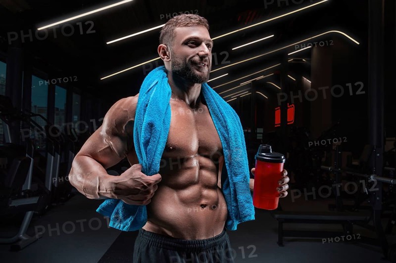 Portrait of a muscular guy with a naked torso and abs. Gym. Blue towel and red shaker. Fitness concept