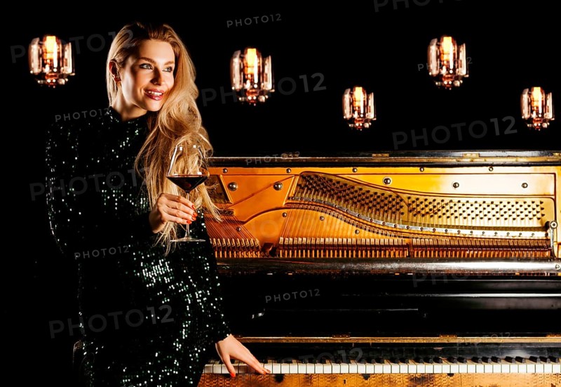 Charming girl in evening dress sits on the piano and poses for the camera with a glass of wine.