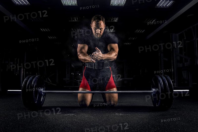 Ahlete kneels in the gym before the bar and claps his hands after which the dust from the chalk flies