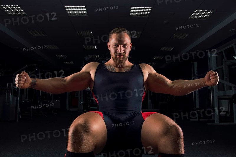 The weightlifter sits on a bench in the gym with arms outstretched and smiles