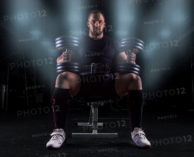Professional weightlifter sits on a bench in the gym with two dumbbells on his lap