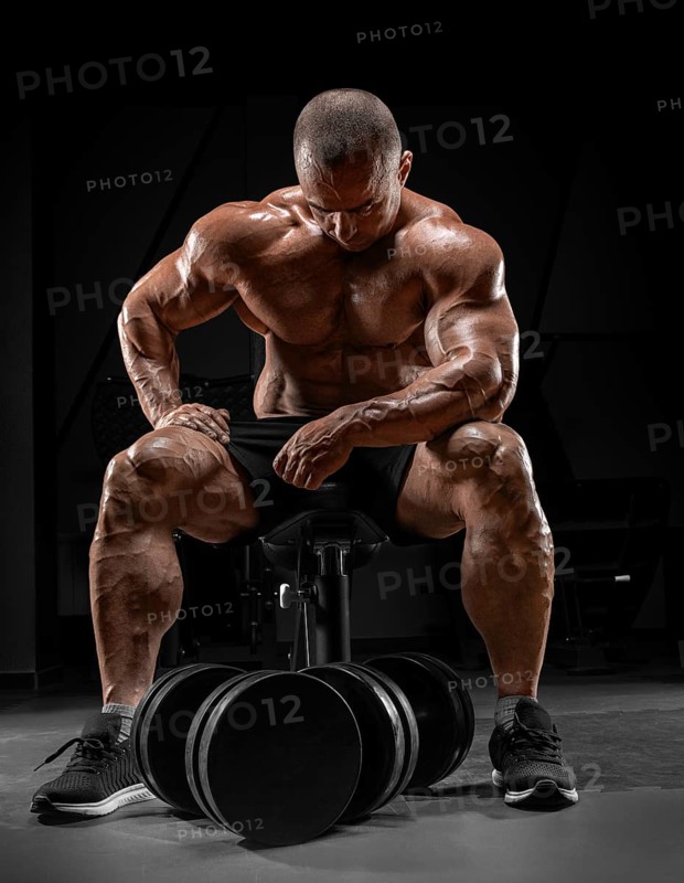 Muscular man sits on a bench near huge dumbbells. Bodybuilding and powerlifting concept.