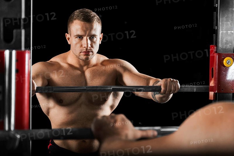 The weightlifter keeps his hands on the barbell and looks at himself in the mirror. Reflection in the Mirror