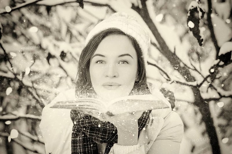 Charming woman blows bright dust from the pages of a book in the winter forest. Fairy tale concept. Christmas. New Year's holidays.