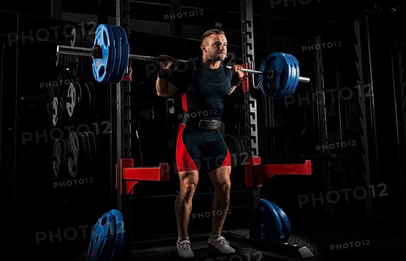The weightlifter stands with a very heavy barbell on his shoulders. He is at the top and is preparing to sit down