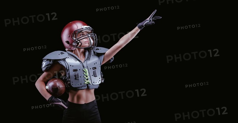 Image of a girl in the uniform of an American football team player. She points to an empty place. Black background. Sports concept. Shoulders pads.