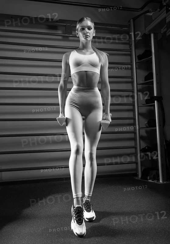 Beautiful tall blonde posing in the gym with dumbbells in her hands against the background of the wall bar. The concept of sports