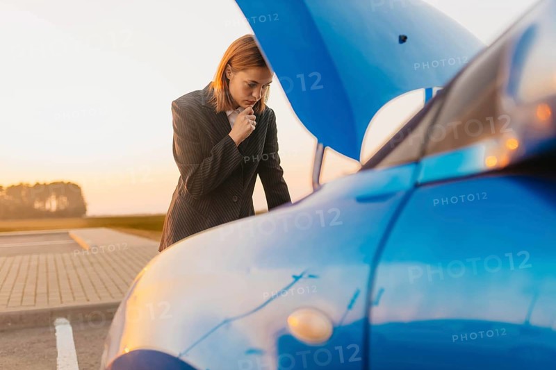 A young beautiful stylish girl driver in a jacket is trying to identify the cause of a car breakdown under the hood