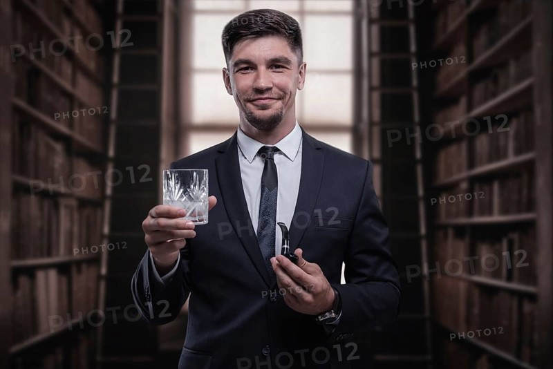 Portrait of an elegant man in a suit with a smoking pipe and a glass in the library. Business concept.