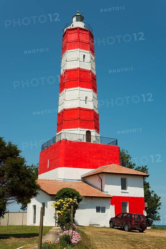 A red and white striped lighthouse next to a house with a vehicle in front of it under a clear sky