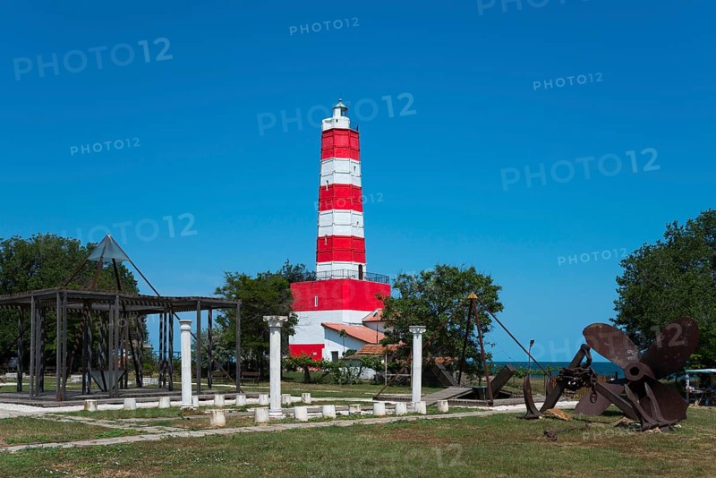Red and white lighthouse under a blue sky next to green areas and a historic anchor