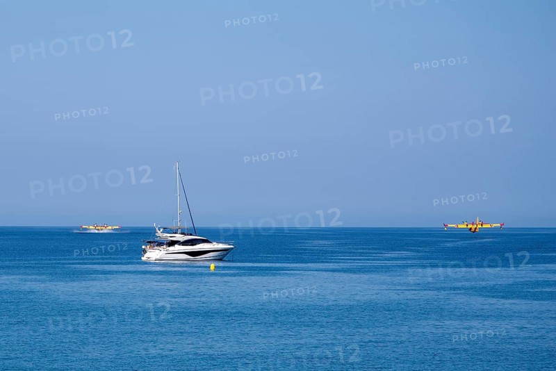 Two fire-fighting aircraft refuelling water at the sea with sailing yacht in the foreground