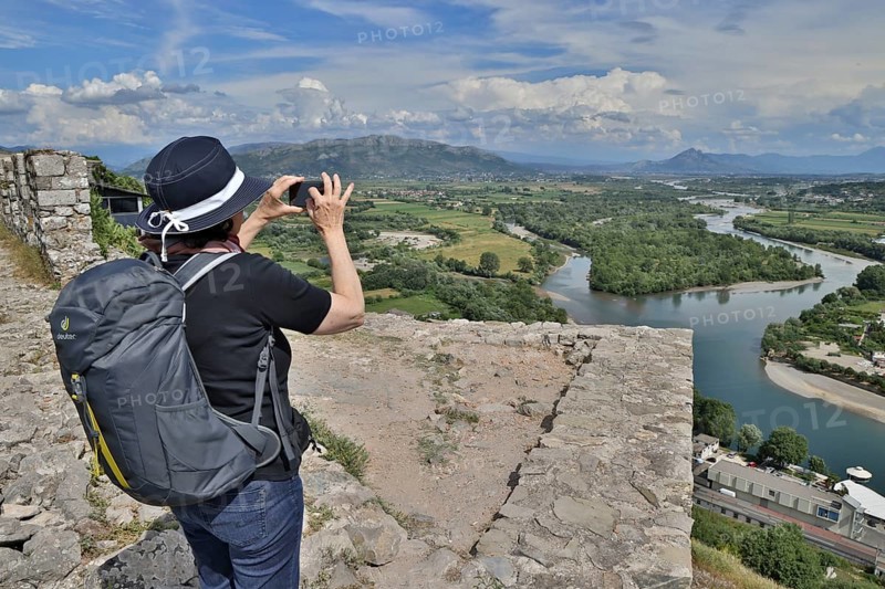 Woman in a sun hat takes a photo with her mobile phone of the view from the Illyrian fortress Rozafa to the river Drin