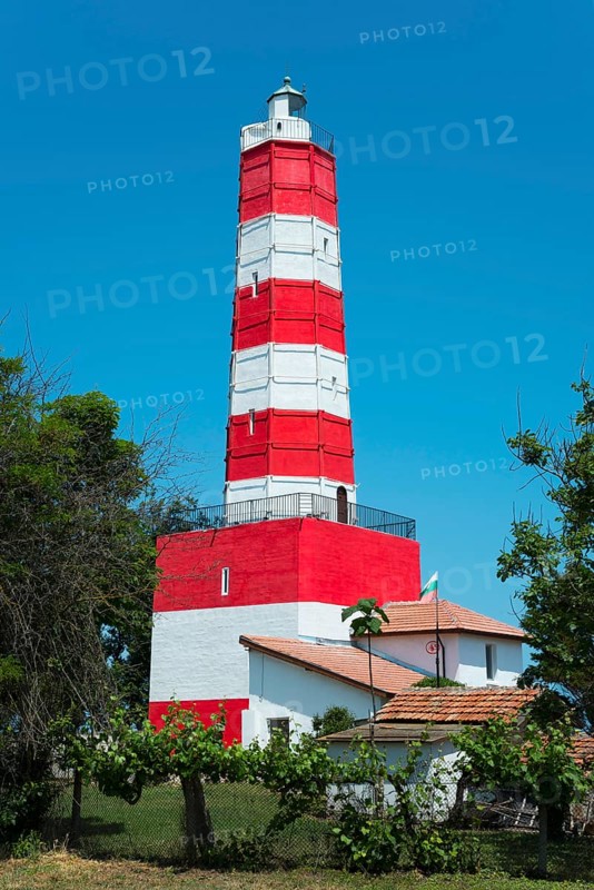 A red and white striped lighthouse stands under a blue sky surrounded by green