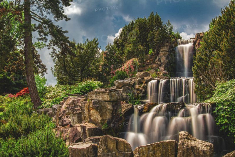 A picturesque waterfall flows over rocky steps surrounded by lush vegetation and dramatic skies, Gruga, Essen, North Rhine-Westphalia, Germany, Europe