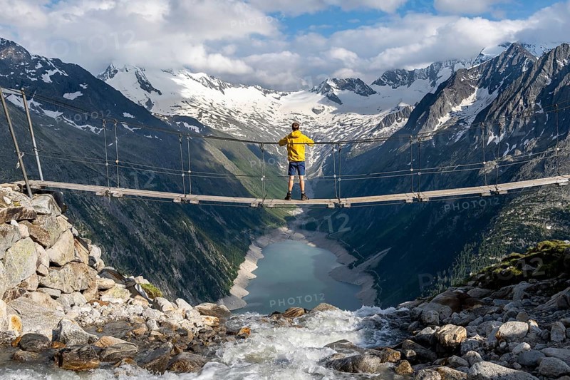 Mountaineers on a suspension bridge over a mountain stream Alelebach, picturesque mountain landscape near the Olpererhuette, view of turquoise-blue lake Schlegeisspeicher, glaciated rocky mountain peaks Hoher Weisszint and Hochfeiler with glacier Schlegeiskees, Berliner Hoehenweg, Zillertal Alps, Tyrol, Austria, Europe