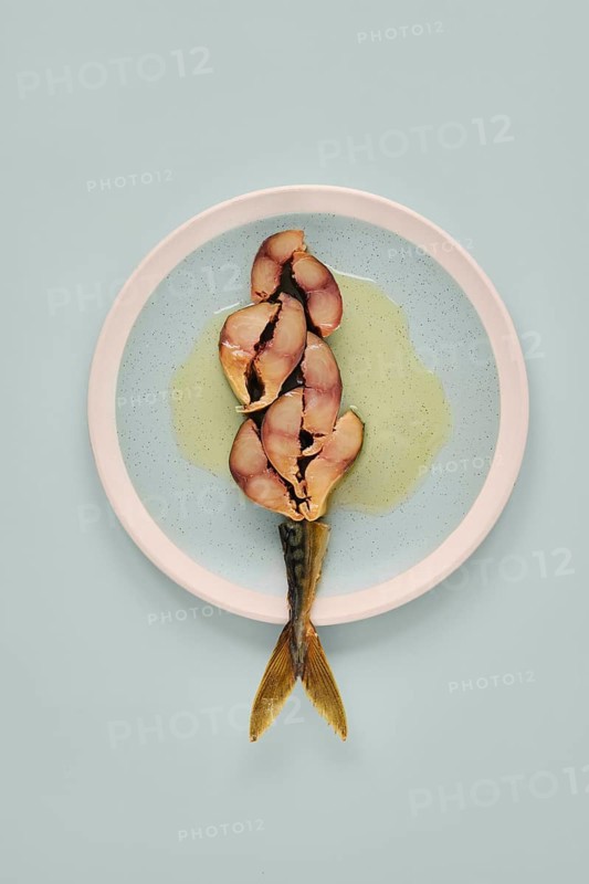Sliced mackerel on a plate revealing its pinkish inner flesh against the darker outer skin. The plate contrasts with both the light blue background and the colors of the fish and sauce