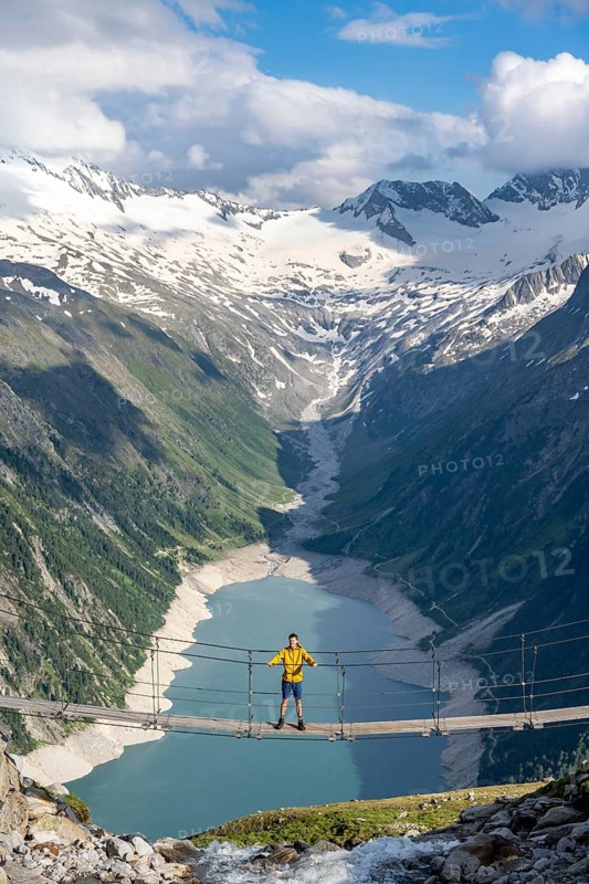 Mountaineers on a suspension bridge over a mountain stream Alelebach, picturesque mountain landscape near the Olpererhuette, view of turquoise-blue lake Schlegeisspeicher, glaciated rocky mountain peaks Grosser Moeseler, Hoher Weisszint and Hochfeilermit glacier Schlegeiskees, Berliner Hoehenweg, Zillertal Alps, Tyrol, Austria, Europe
