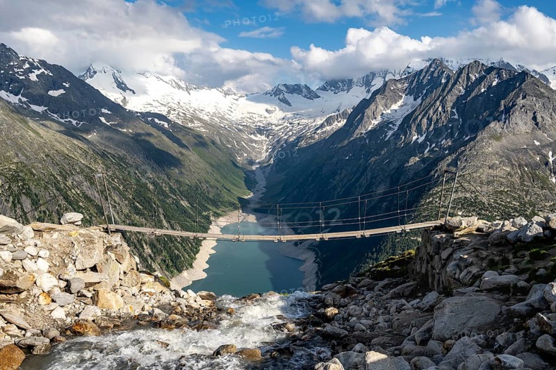 Suspension bridge over a mountain stream Alelebach, picturesque mountain landscape near the Olpererhuette, view of turquoise-blue lake Schlegeisspeicher, glaciated rocky mountain peaks Grosser Moeseler, Hoher Weisszint and Hochfeilermit glacier Schlegeiskees, Berliner Hoehenweg, Zillertal Alps, Tyrol, Austria, Europe