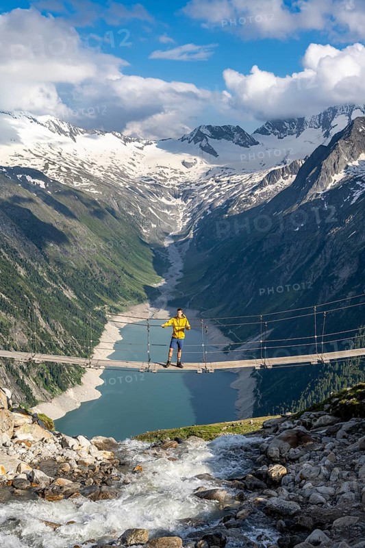 Mountaineers on a suspension bridge over a mountain stream Alelebach, picturesque mountain landscape near the Olpererhuette, view of turquoise-blue lake Schlegeisspeicher, glaciated rocky mountain peaks Grosser Moeseler, Hoher Weisszint and Hochfeilermit glacier Schlegeiskees, Berliner Hoehenweg, Zillertal Alps, Tyrol, Austria, Europe