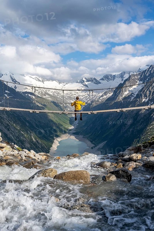 Mountaineer sitting on a suspension bridge over a mountain stream Alelebach, picturesque mountain landscape near the Olpererhuette, view of turquoise blue lake Schlegeisspeicher, glaciated rocky mountain peaks Hoher Weisszint and Hochfeiler with glacier Schlegeiskees, Berliner Hoehenweg, Zillertal Alps, Tyrol, Austria, Europe