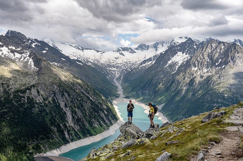 Two mountaineers on a rock in front of a mountain panorama, view of Schlegeisspeicher, glaciated rocky mountain peaks Hoher Weisszint and Hochfeiler with glacier Schlegeiskees, Berliner Hoehenweg, Zillertal Alps, Tyrol, Austria, Europe