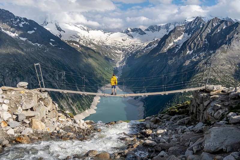 Mountaineers on a suspension bridge over a mountain stream Alelebach, picturesque mountain landscape near the Olpererhuette, view of turquoise-blue lake Schlegeisspeicher, glaciated rocky mountain peaks Hoher Weisszint and Hochfeiler with glacier Schlegeiskees, Berliner Hoehenweg, Zillertal Alps, Tyrol, Austria, Europe