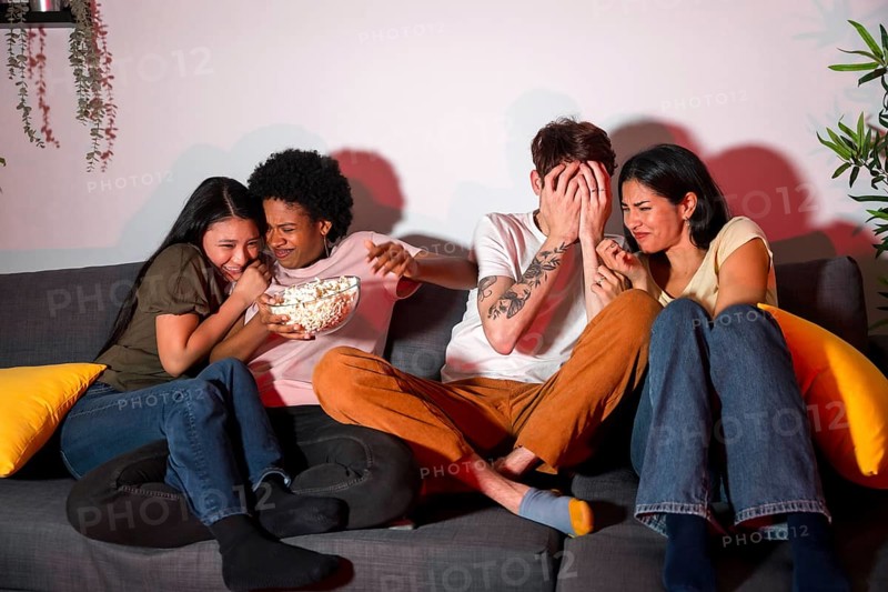 Multi-ethnic friends on a terror movie night at home sitting on the sofa eating popcorn and covering faces