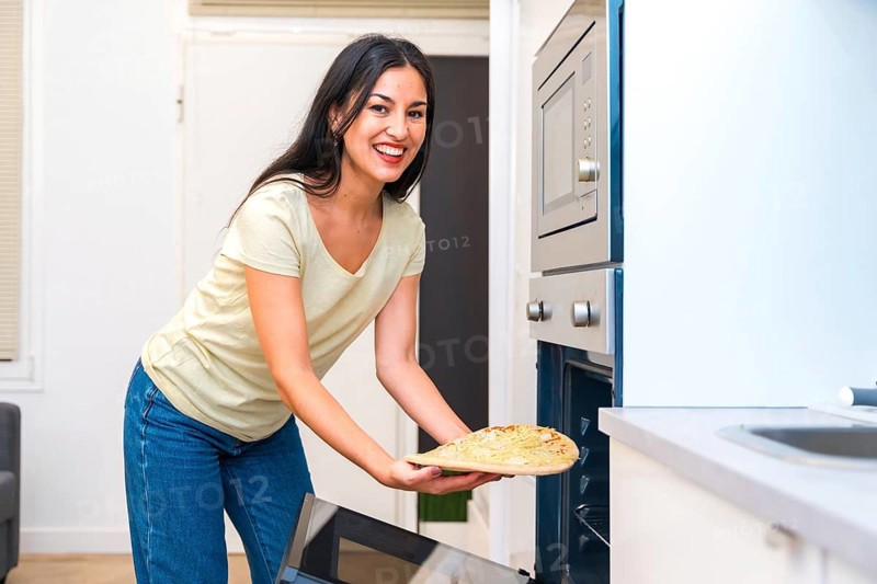 Smiling woman looking at camera while putting a pizza into a oven at home