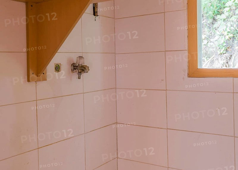 Water faucet on white tiled wall next to wooden shelf and window in laundry room of abandoned house