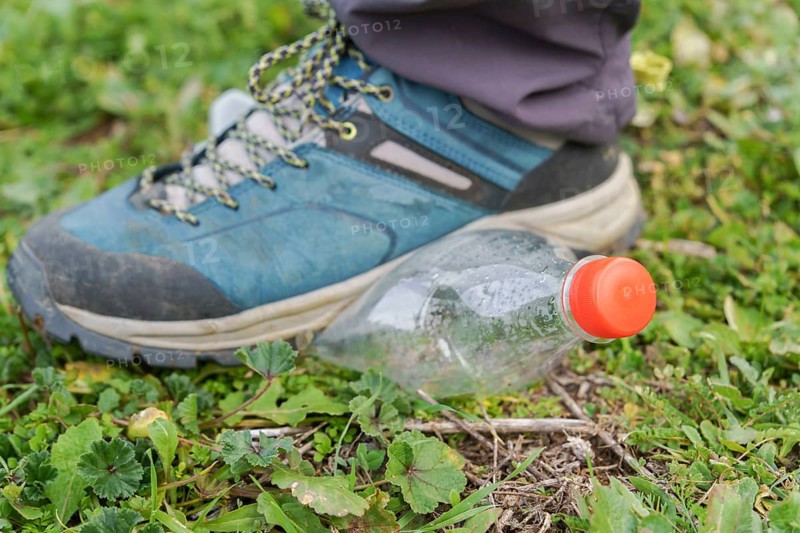 Hiking boot stepping on an empty plastic bottle on the green grass of a meadow, ecology and environment concept