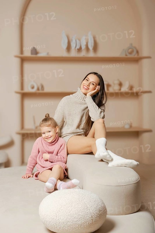 Happy young mother in warm sweater and socks sitting on top of a white ottoman next to her daughter