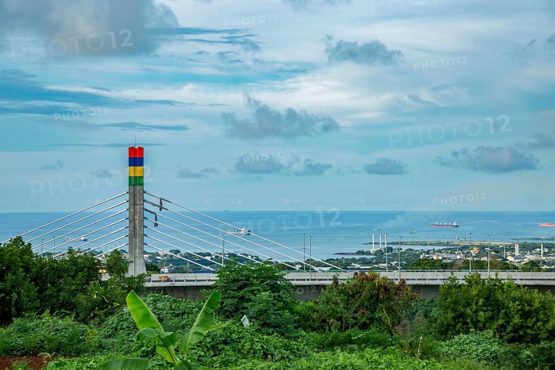 A highway with Mauritian emblem on pillar and a truck driving, surrounded by mountains and an overcast sky.A1-M1 Link Bridge At Grand River North West Valley on the island of Mauritius