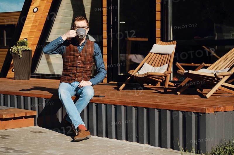 Middle-aged man sitting on terrace of wooden cabin and drinking tea in the morning
