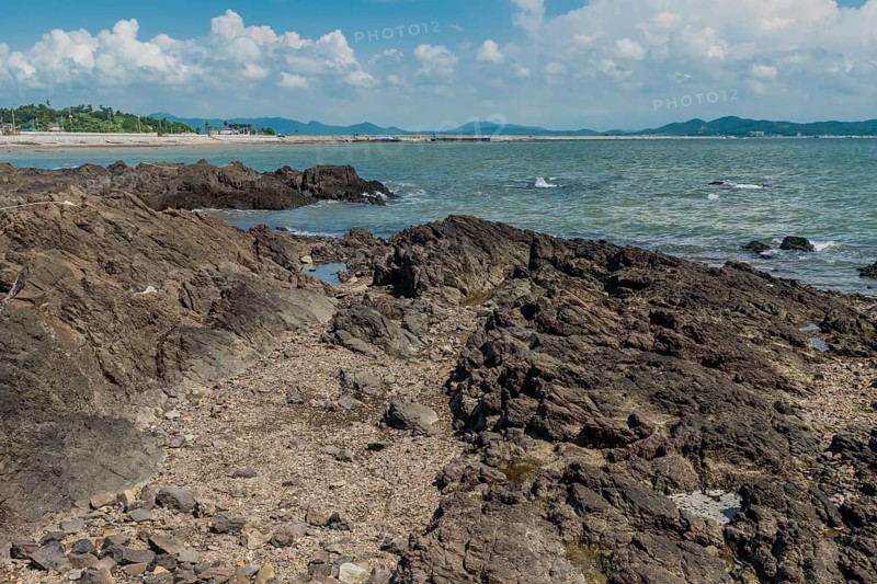 Seascape of rocking shore with beach and harbor road in background under cloudy blue sky