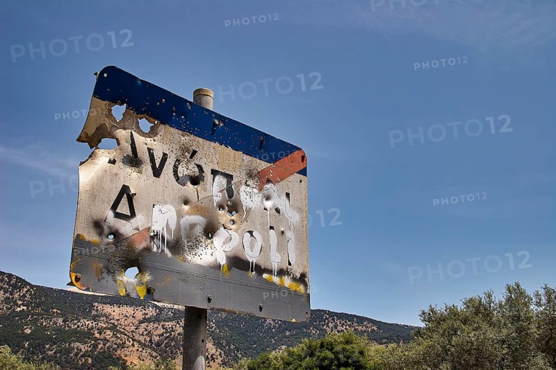 Weathered road sign with bullet holes against a blue sky and trees in the background, Anopolis, Sfakia, West Crete, Crete, Greece, Europe