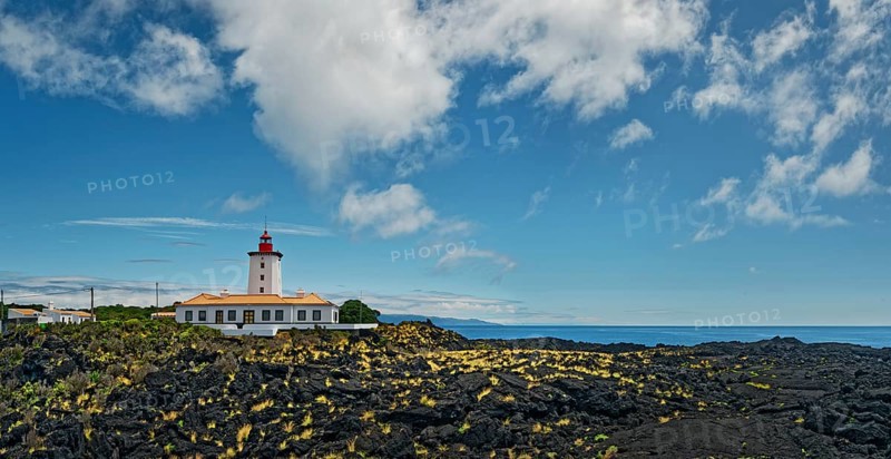 Wide lava flow with the striking lighthouse Farol da Ponta da Iha in front of a blue sky with white clouds, lava rocks coastal path Ponta da Iiha, Manhenha, west coast, Pico, Azores