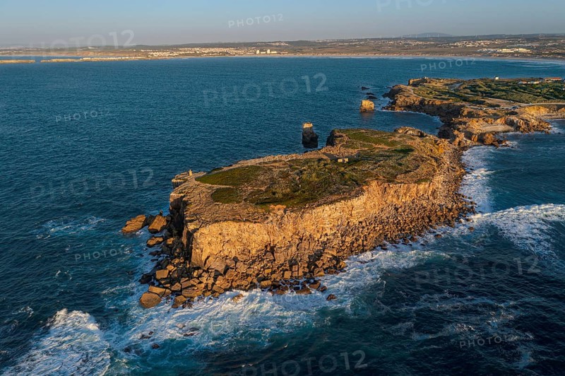 Aerial image of high rocky headland and peninsula. Drone shot of sea blue waves beat and splash in summer sunset haze, little foliage and rocky cliffs, establishing or static shot, Peniche, Portugal, Europe