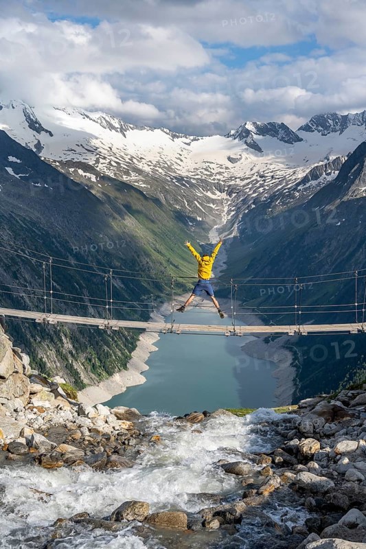 Mountaineer jumping on a suspension bridge over a mountain stream Alelebach, picturesque mountain landscape near the Olpererhuette, view of turquoise blue lake Schlegeisspeicher, glaciated rocky mountain peaks Hoher Weisszint and Hochfeiler with glacier Schlegeiskees, Berliner Hoehenweg, Zillertal Alps, Tyrol, Austria, Europe