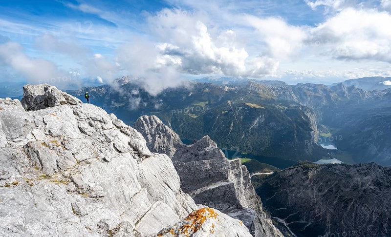 Mountaineer on the rocky summit of the Watzmann Mittelspitze, Watzmann crossing, view of mountain panorama with Steinernes Meer and Koenigssee, Berchtesgaden National Park, Berchtesgaden Alps, Bavaria, Germany, Europe