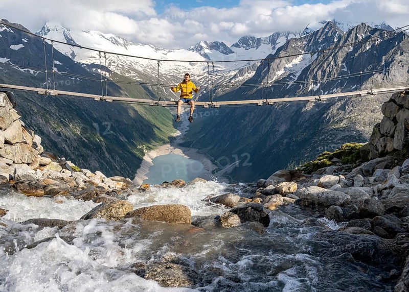 Mountaineer sitting on a suspension bridge over a mountain stream Alelebach, picturesque mountain landscape near the Olpererhuette, view of turquoise blue lake Schlegeisspeicher, glaciated rocky mountain peaks Hoher Weisszint and Hochfeiler with glacier Schlegeiskees, Berliner Hoehenweg, Zillertal Alps, Tyrol, Austria, Europe