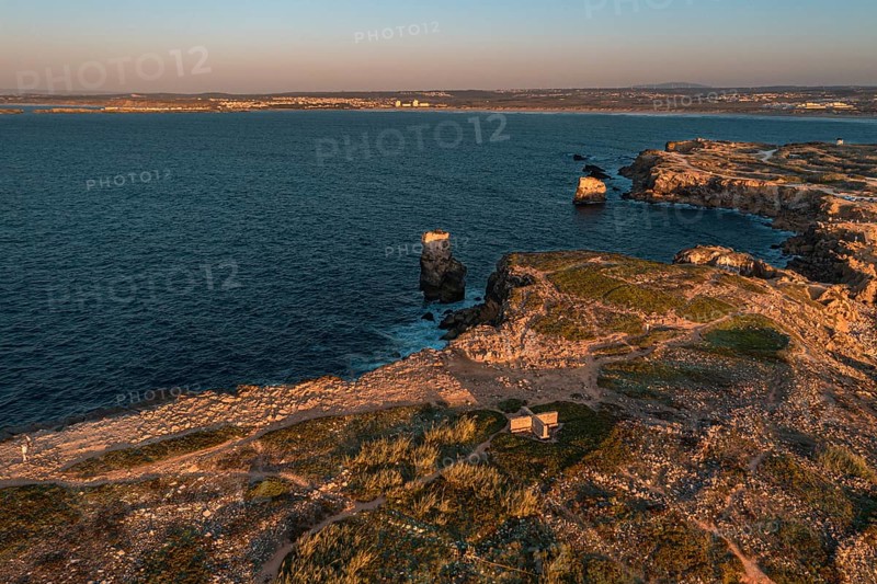 Bird's-eye view of sunset over ocean waves break on rocky shores, Peniche, Portugal. Summer sunset haze, little foliage and rocky cliffs, peninsula and rocks, fishing town, horizon