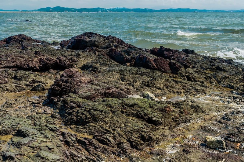 Seascape of rocking shore with beach and harbor road in background under cloudy blue sky