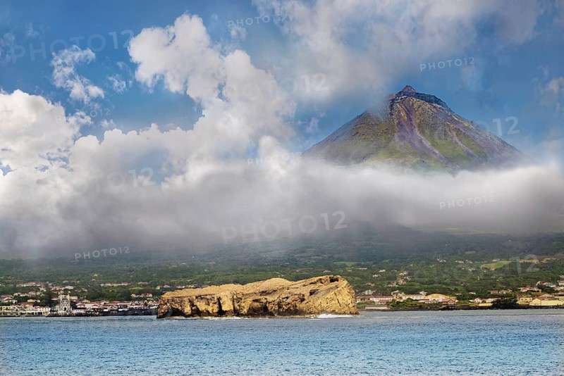 The volcano Pico rises from behind a cloud cover, surrounded by the blue sea with the rocks of Iieu Deitado and the town of Madalena, Iieu Deitado, Horta, Faial, Azores, Portugal, Europe