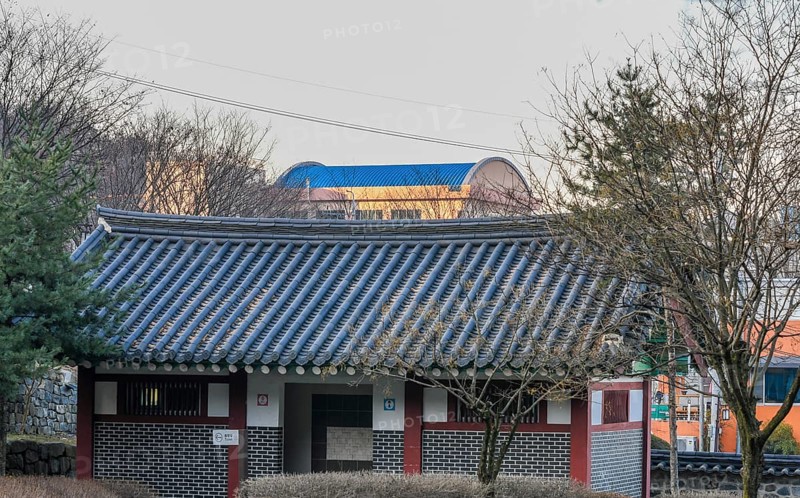 Public restroom in woodland park with a tiled roof surrounded by trees and brushes with a modern building with a blue roof in the background