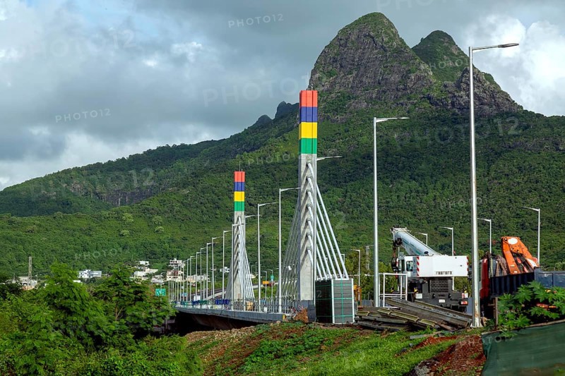 A highway with Mauritian emblem on pillar and a truck driving, surrounded by mountains and an overcast sky.A1-M1 Link Bridge At Grand River North West Valley on the island of Mauritius