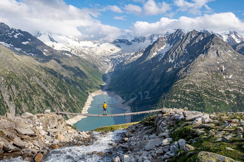 Mountaineers on a suspension bridge over a mountain stream Alelebach, picturesque mountain landscape near the Olpererhuette, view of turquoise-blue lake Schlegeisspeicher, glaciated rocky mountain peaks Grosser Moeseler, Hoher Weisszint and Hochfeiler with glacier Schlegeiskees, Berliner Hoehenweg, Zillertal Alps, Tyrol, Austria, Europe