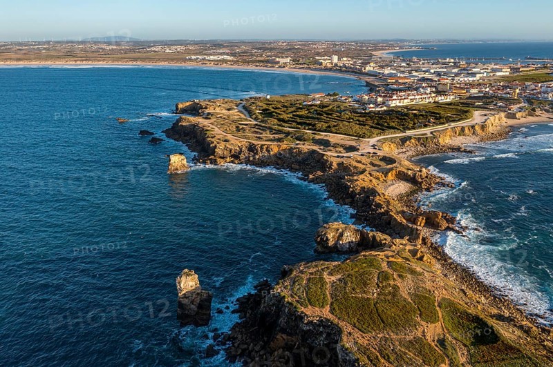 Aerial image of peninsula and town of Peniche, Portugal on sunny day. Summer sunset haze, little foliage and rocky cliffs, peninsula and rocks, fishing town, horizon, ocean waves break on rocky shores