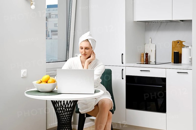 Serious young woman in bathrobe sits in the kitchen with laptop