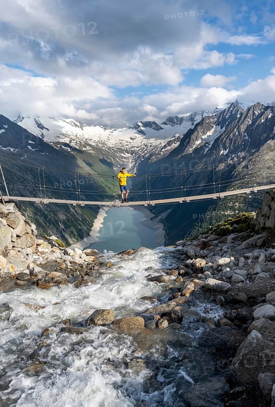 Mountaineers on a suspension bridge over a mountain stream Alelebach, picturesque mountain landscape near the Olpererhuette, view of turquoise-blue lake Schlegeisspeicher, glaciated rocky mountain peaks Hoher Weisszint and Hochfeiler with glacier Schlegeiskees, Berliner Hoehenweg, Zillertal Alps, Tyrol, Austria, Europe